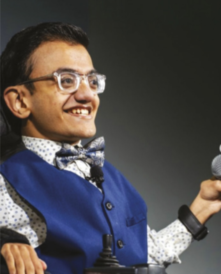 Fotografia de Sparsh Shah sorrindo, segurando um microfone. Ele é branco, tem cabelos curtos, lisos e castanhos, usa óculos e está de camisa, colete e gravata borboleta. Seus braços e tronco são curtos e ele está em uma cadeira de rodas motorizada.
