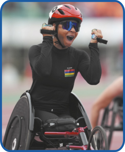 Fotografia de Noemi Alphonse comemorando enquanto corre em uma pista de atletismo em uma cadeira de rodas. Ela é parda, tem as pernas amputadas e está usando capacete e blusa de manga longa.
