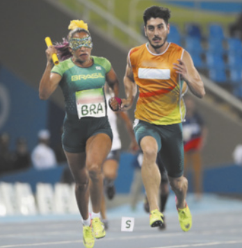 Fotografia de Terezinha Guilhermina durante uma prova de atletismo paralímpico. A atleta brasileira usa uniforme verde com a inscrição 'BRA' e corre segurando um bastão de revezamento. Ao lado dela, um atleta-guia de pele clara, com uniforme nas cores laranja e verde, a acompanha na pista. Ambos estão em movimento, com a arquibancada azul desfocada ao fundo.