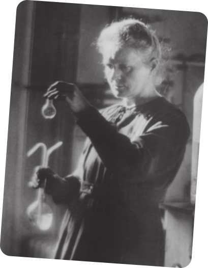 Foto em preto e branco de Marie Curie em pé, manipulando balões volumétricos em seu laboratório. Ela está usando vestido de mangas longas.
