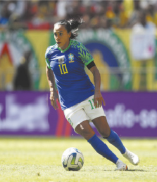 Fotografia de Marta Silva durante uma partida de futebol. Ela tem cabelos pretos presos para trás e veste o uniforme da seleção brasileira: camiseta azul e shorts branco, meia azul e chuteira branca. Ao fundo uma arquibancada desfocada.
