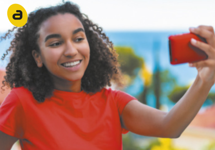 A. Fotografia de uma jovem sorridente segurando um celular com uma das mãos, esticado na altura do rosto, tirando uma selfie. Ela tem cabelos curtos e crespos, e veste uma camiseta vermelha.