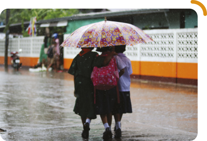 Fotografia de três pessoas caminhando em uma rua com o chão molhado. Elas estão de costas, próximas umas das outras, e compartilham um guarda-chuva grande e colorido, com estampa de flores. Ao fundo, há um muro branco com faixa laranja e algumas árvores.