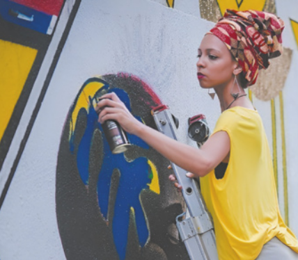 Fotografia de Criola pintando um mural com tinta spray. Ela está no topo de uma escada encostada em um muro. Está pintando manchas amarelas e azuis sobre a imagem da cabeça de um homem negro. Criola usa um turbante estampado cobrindo os cabelos, brincos longos e camiseta.
