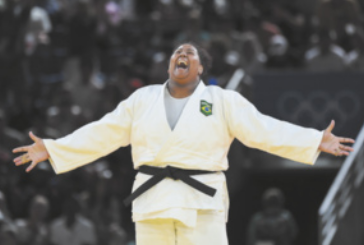 Fotografia da judoca Beatriz Souza com os braços abertos, sorrindo com a cabeça levantada. Ela é negra, tem cabelos curtos e crespos, usa um quimono branco com uma faixa preta amarrada na cintura e está em um ginásio, com público nas arquibancadas ao fundo.