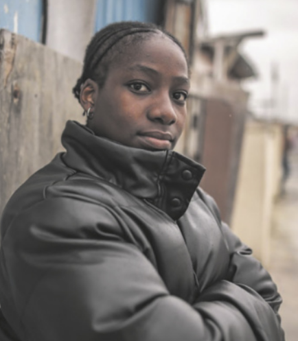 Fotografia de Yola Mgogwana com expressão séria e os braços cruzados. Ela é negra, tem os cabelos crespos trançados na raiz e usa uma jaqueta de frio. Ela está na rua, à frente de tapumes.