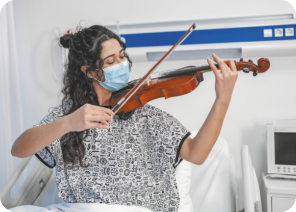 Fotografia de uma jovem sentada na cama de um hospital, tocando violino. Ela é branca, tem cabelos longos, pretos e ondulados, e está usando máscara de proteção e roupa hospitalar estampada.