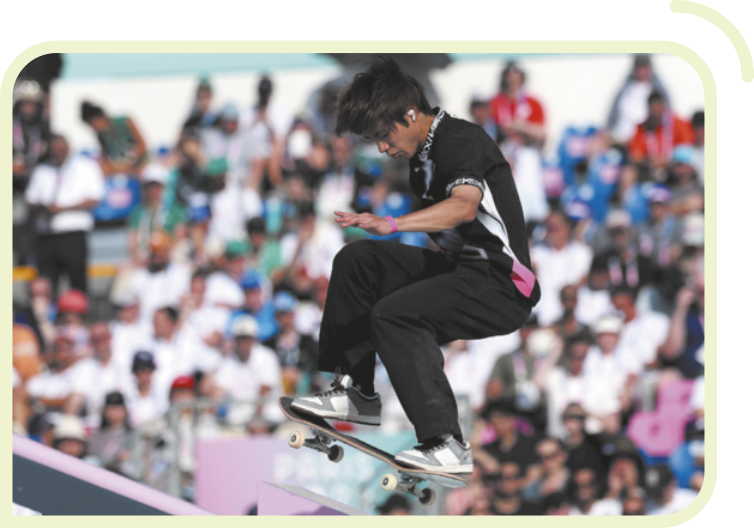 Fotografia de Yuto Horigami saltando de skate. Ele tem traços asiáticos, cabelos curtos e lisos e está usando camiseta, calça e tênis. O público observa das arquibancadas, ao fundo.