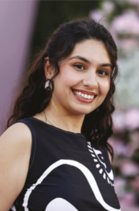 Fotografia de Alessia Cara sorrindo. Mulher de pele branca e cabelos castanhos escuros, longos e cacheados, presos parcialmente para trás. Ela usa um vestido preto com estampa branca e brincos. Ao fundo, há uma parede com tons claros, desfocada.