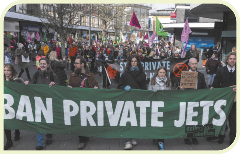 Fotografia de uma manifestação na rua. À frente, homens e mulheres seguram uma faixa, onde se lê: 'BAN PRIVATE JETS'. Ao fundo, muitas outras pessoas caminham, segurando bandeiras e outras faixas.