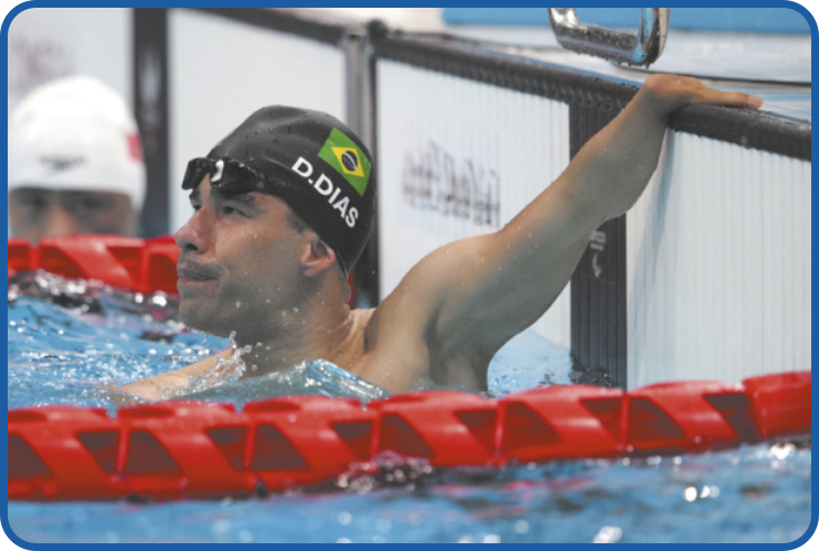 Fotografia de Daniel Dias em uma piscina. Ele usa óculos de natação e uma touca preta com a bandeira do Brasil e seu nome, 'D. Dias', estampados. É possível observar a má formação congênita no braço que ele está apoiando na borda da piscina.