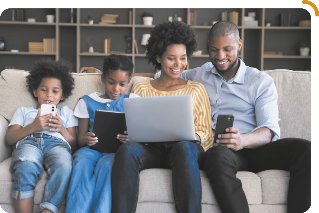 Fotografia de uma família sentada no sofá, cada um utilizando um equipamento eletrônico. O menino usa um celular, a menina, um tablet, a mulher está com um notebook apoiado no colo, enquanto observa a tela do celular na mão do homem, que também está olhando para a tela.
