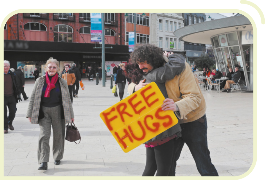 Fotografia de um homem abraçando uma pessoa na rua. Ele segura um cartaz amarelo com a frase 'Free Hugs' escrita em letras vermelhas. Ao fundo, aparecem outras pessoas andando, prédios com janelas e fachadas comerciais.