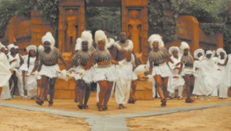 Fotografia de uma cena do filme 'Black Panther'. Em primeiro plano, um grupo de dançarinos realiza uma apresentação, vestindo roupas brancas, blusas e adornos na cabeça. A cena acontece em um espaço ao ar livre, com uma construção com colunas e esculturas, rodeada por vegetação.