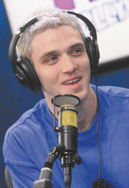 Fotografia de Lauv falando ao microfone em um estúdio. Ele está sentado, usa fones de ouvido, é um jovem branco com os cabelos curtos e descoloridos.