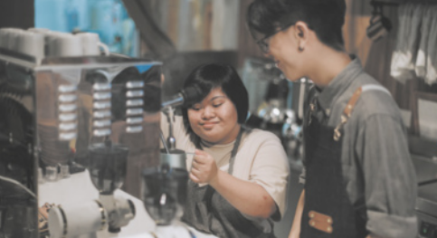 Fotografia de um jovem e uma jovem trabalhando em uma cafeteria. Eles têm traços asiáticos e estão usando avental. A garota tem cabelos curtos e lisos na altura da nuca e está com um bule de inox em um dos bicos de uma máquina de café expresso. O garoto a observa, ao lado.