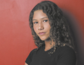 Fotografia de Catarina Lorenzo em frente a uma parede vermelha. Ela é uma jovem branca, tem cabelos castanhos, ondulados e longos, e está usando uma camiseta preta.