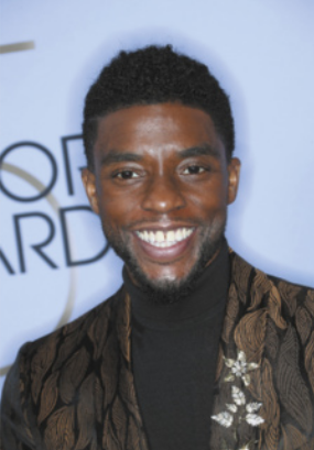 Fotografia de Chadwick Boseman. Ele está de pé, tem cabelo curto e crespo, usa uma blusa de gola alta preta e um paletó marrom com um broche. Ao fundo, há um painel branco com letras em tom escuro.