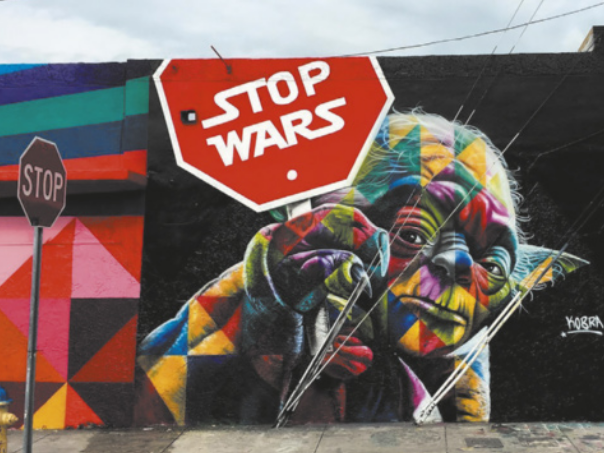 Fotografia de um mural de Eduardo Kobra. A imagem mostra um Jedi de Star Wars segurando uma placa vermelha com os dizeres em branco 'Stop Wars'. O mural é colorido, com traços geométricos e vibrantes. Na calçada, há uma placa de trânsito com a palavra 'STOP'.