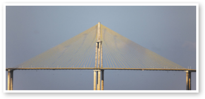 Fotografia de uma ponte com formato simétrico. No centro há um mastro alto, de onde partem vários cabos finos que descem em direção ao tabuleiro da ponte, formando uma estrutura triangular.