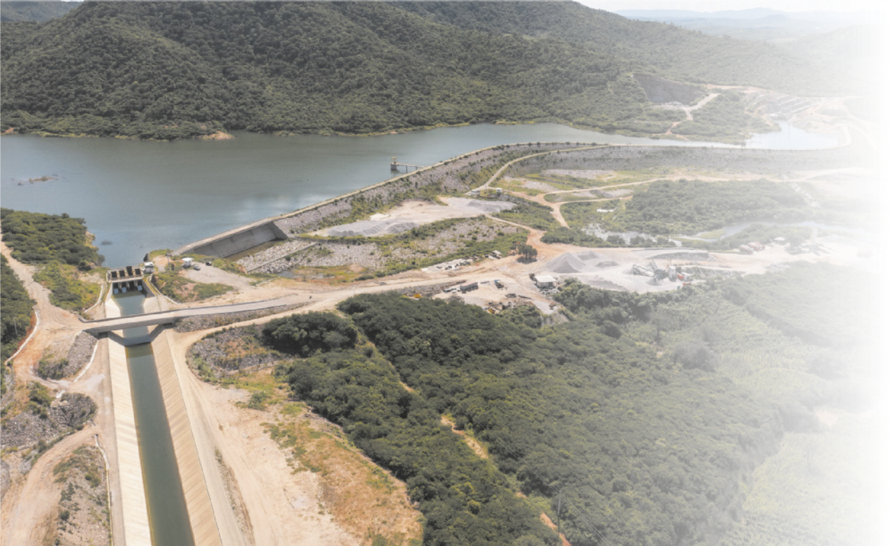 Fotografia aérea de um açude. É possível observar o reservatório de água, a barragem e a área operacional. Há morros com vegetação ao redor.