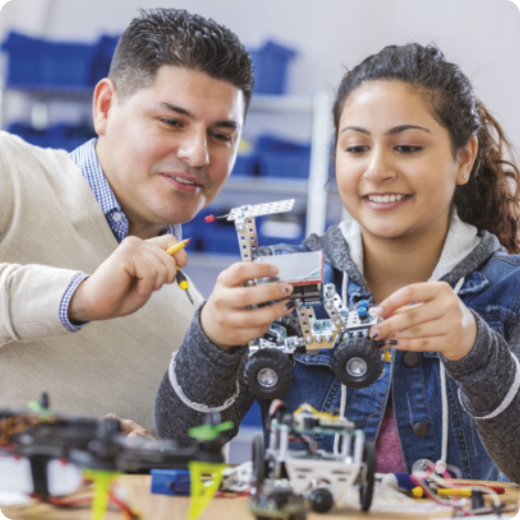 Fotografia de um homem adulto e uma jovem montando juntos uma estrutura que se assemelha a uma escavadeira em miniatura, feita com peças de robótica.