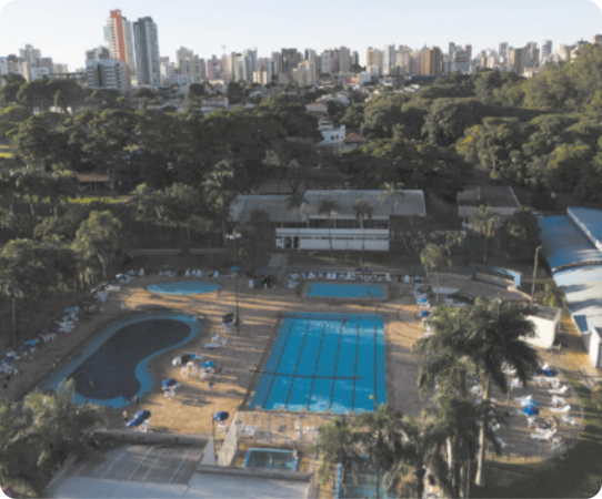 Fotografia aérea de um clube. É possível observar duas piscinas adultas e duas infantis, com várias espreguiçadeiras ao redor. Há vegetação no entorno.