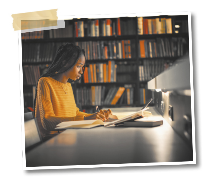 Fotografia de uma mulher sentada à mesa em uma biblioteca, utilizando um notebook e fazendo anotações.
