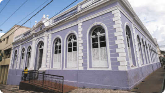 Fotografia de um prédio antigo em forma de bloco retangular, com grandes janelas em forma de arco ao longo da fachada. Há uma rampa de acesso na entrada.