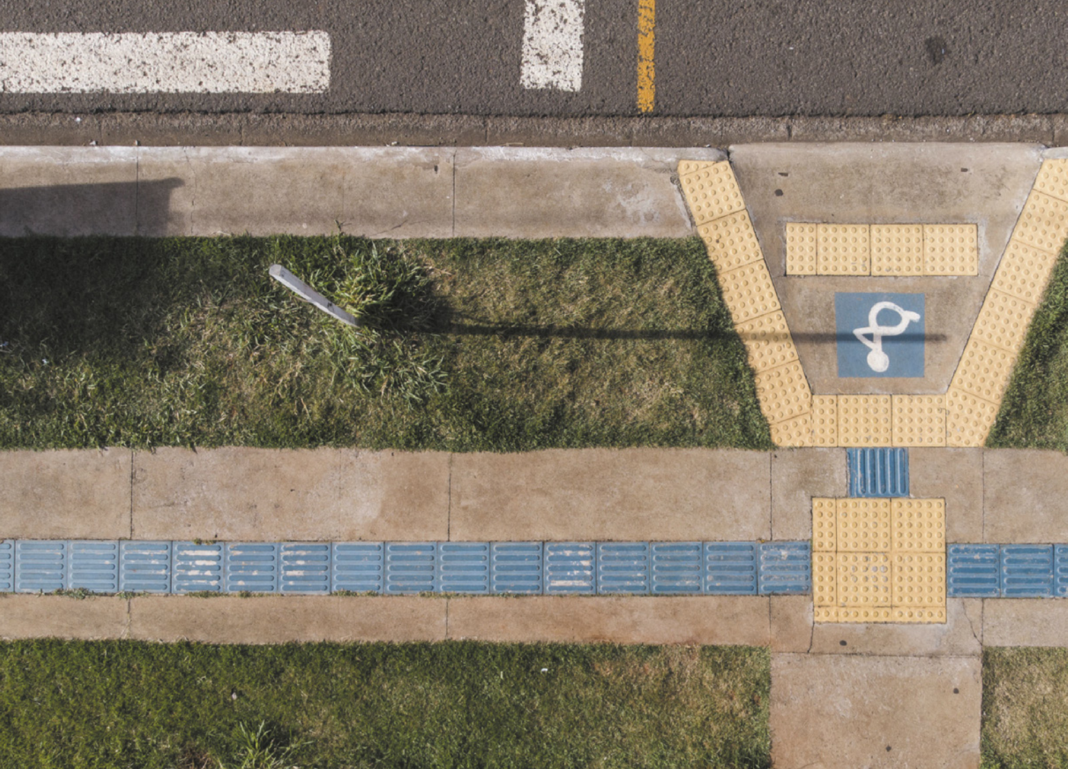 Fotografia aérea de uma calçada. É possível observar uma rampa de acesso para cadeirantes, que leva até a rua. Há piso tátil com tiras retas na cor azul, que percorrem longitudinalmente a calçada, além de piso tátil com bolinhas na cor amarela, presente antes da rampa de acesso e também em suas extremidades.