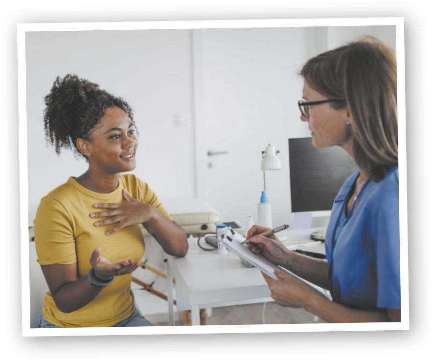 Imagem de um ambiente interno de consultório. À esquerda, uma mulher jovem gesticula com uma das mãos sobre o peito enquanto conversa com uma profissional de saúde. À direita, a profissional, de óculos e uniforme, escuta com atenção e faz anotações em uma prancheta.