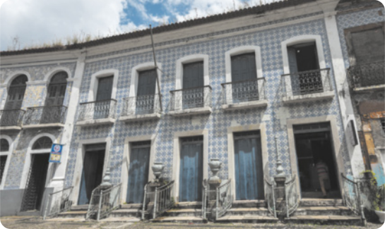 Imagem de uma fachada de construção antiga, com dois andares e revestida de azulejos. O prédio tem estilo colonial, com 7 portas no pavimento térreo e 7 no andar superior, acompanhadas por sacadas com grade de ferro. A fachada está desgastada, com vegetação crescendo no topo do edifício.