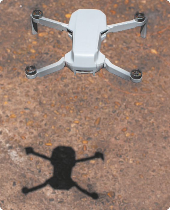 Imagem de um drone visto de cima, pairando sobre um solo de terra batida. Abaixo do drone, sua sombra é projetada no chão, com o mesmo formato do corpo e das quatro hélices, dispostas duas em cada lado.