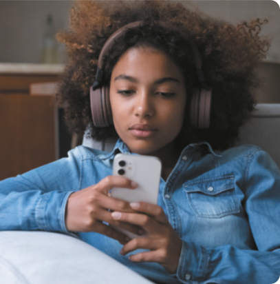Fotografia de uma jovem sentada em um sofá, usando fones de ouvido e olhando para o celular que segura com as duas mãos. Ela veste uma camisa jeans e tem cabelos cacheados e volumosos. Sua expressão é neutra e concentrada.