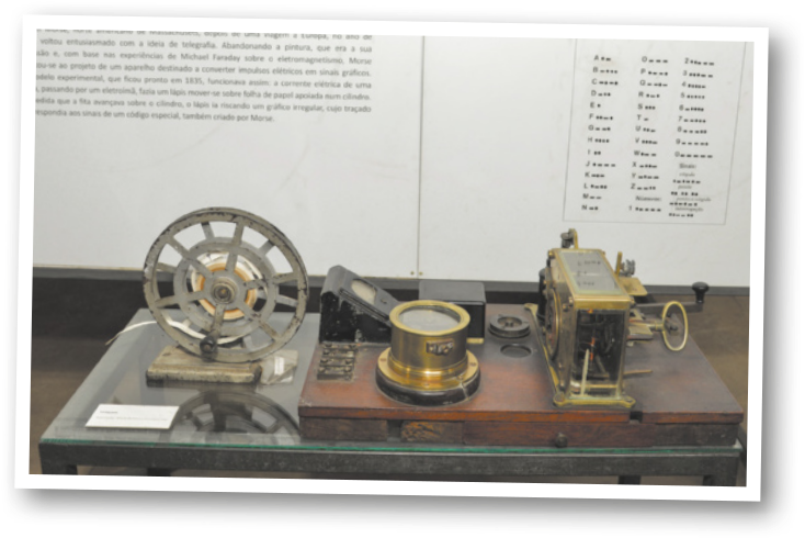 Imagem de dois equipamentos antigos expostos sobre uma mesa. Ao fundo, há um painel com texto explicativo e uma tabela do código Morse.