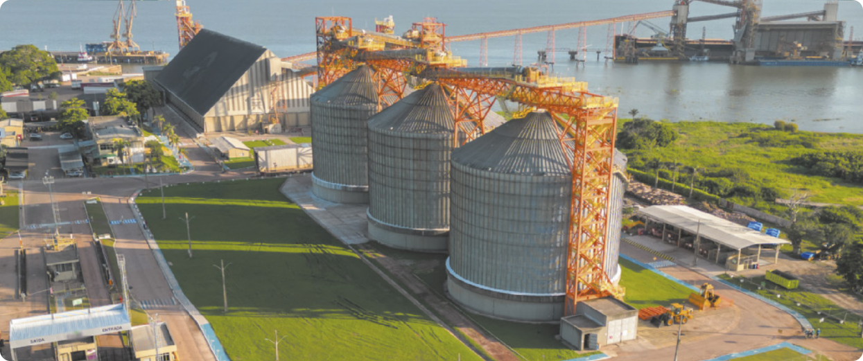 Imagem aérea de uma área industrial próxima a um corpo d’água, com destaque para três silos metálicos de grande porte. Cada silo tem a forma de um cilindro com cobertura cônica. Ao redor, há áreas com gramados, construções menores e vias de acesso pavimentadas.