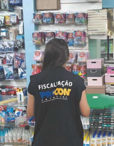 Fotografia de uma mulher, vista de costas, em uma loja de artigos de papelaria. Ela veste um colete preto com a inscrição 'Fiscalização PROCON'. Ao fundo, há caixas organizadoras empilhadas e diversos produtos escolares, como estojos, colas e outros materiais.