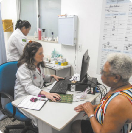 Imagem de uma sala de atendimento médico. À esquerda, uma profissional de saúde usa jaleco e está sentada diante de um computador, conversando com uma paciente idosa. Ao fundo, outra profissional, também de jaleco, organiza materiais sobre uma bancada.