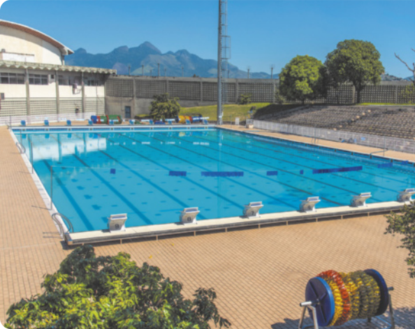 Imagem de uma piscina olímpica ao ar livre, vista em perspectiva. A piscina é retangular, com raias divididas por boias flutuantes, e o piso ao redor é de cor clara. Ao fundo, há arquibancada, grades e algumas árvores.