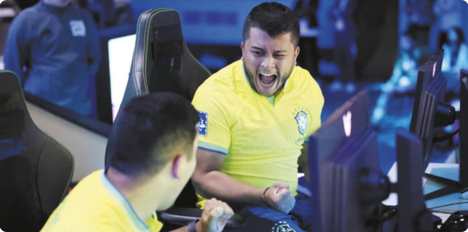 Fotografia de dois homens em frente a computadores. Ambos vestem camisas com o escudo do Brasil. Um deles aparece de perfil, enquanto o outro está de frente, com os punhos cerrados e expressão de euforia. Eles estão sentados em cadeiras 'gamer', cercados por monitores, em um ambiente com iluminação azul.