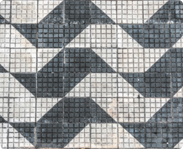 Imagem de ladrilhos quadrados, organizados em um padrão geométrico. Há três tipos de ladrilhos: totalmente brancos, totalmente pretos e ladrilhos divididos diagonalmente em duas metades, uma de cada cor. Esses ladrilhos estão dispostos de modo a formar padrões que se repetem ao longo de toda a imagem. Cada padrão é composto por 8 ladrilhos, dispostos em 2 linhas com 4. Primeira linha: branco, meio a meio, preto, meio a meio. Os ladrilhos meio a meio estão orientados de modo que, com o ladrilho preto, formam um trapézio. Segunda linha: meio a meio, preto, meio a meio e branco. Essa linha repete o padrão do trapézio, mas invertido e deslocado uma posição à esquerda em relação ao da linha superior. A alternância entre essas linhas cria uma sequência de trapézios pretos e brancos que se repetem de forma contínua por toda a imagem.