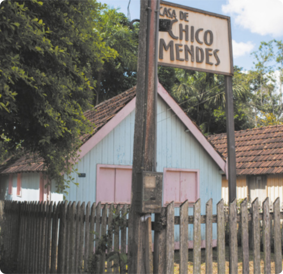 Fotografia de uma casa de madeira, pequena. Na frente dela há uma placa com o escrito Casa de Chico Mendes.