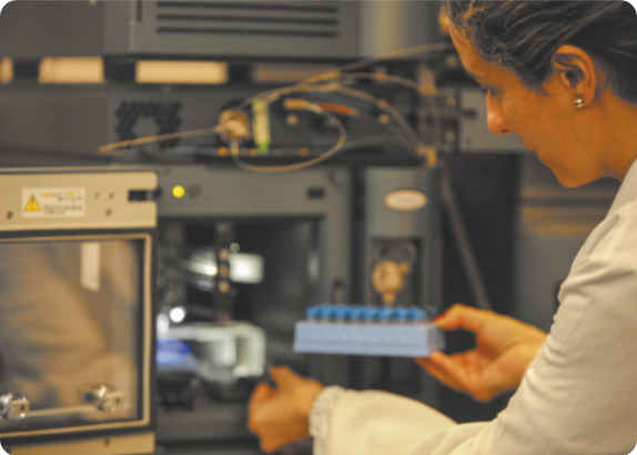 Fotografia de uma mulher em um laboratório. Ela segura um suporte com tubo em frente a um equipamento aberto.