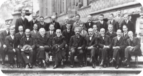 Fotografia de homens formando fileiras. Eles vestem calças e ternos. Entre eles está Albert Einstein, um homem de cabelo grisalho, curto e com bigode.