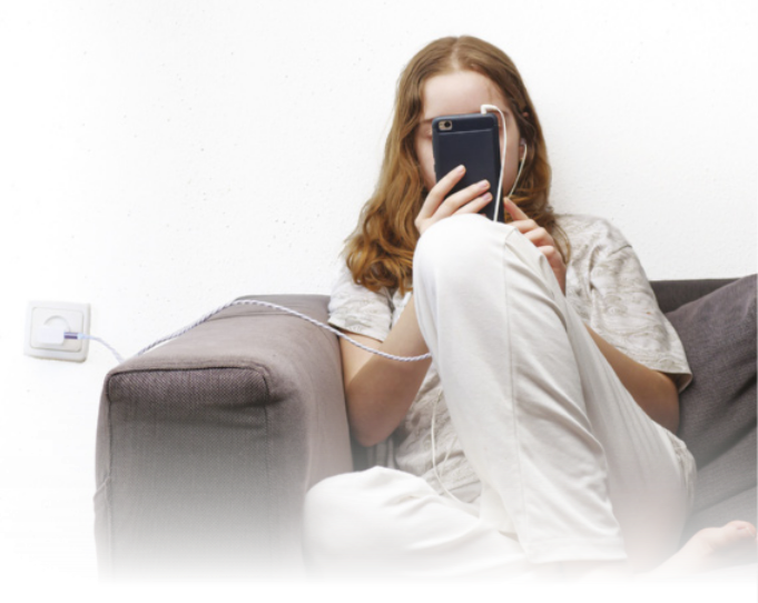 Fotografia de uma jovem sentada ao sofá, utilizando um celular, que está conectado ao carregador.