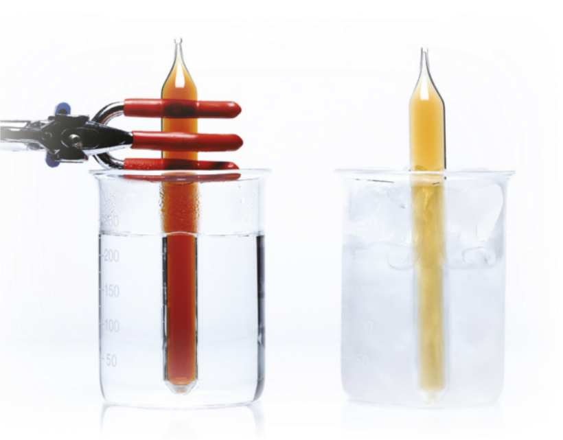 Fotografia de um experimento, em dois momentos. No primeiro, está sendo colocado um frasco fechado com um líquido castanho-avermelhado em um recipiente com água. No segundo, a água do recipiente está congelada e o líquido do frasco fechado agora está amarelo-claro.