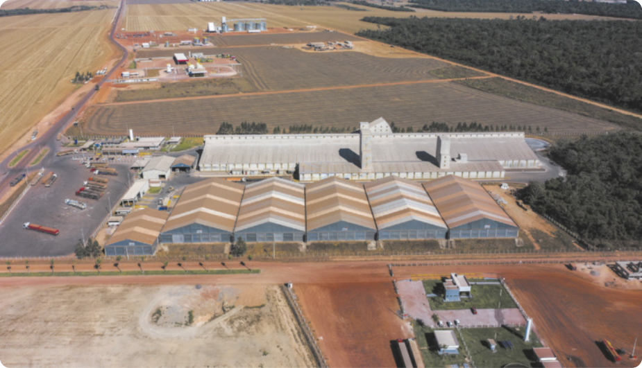 Fotografia aérea de uma fábrica. Há galpões alinhados e outras construções localizadas em uma área rural, próxima a plantações.