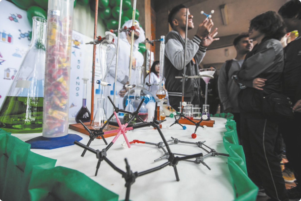 Fotografia de uma mesa com modelos científicos e equipamentos de laboratório. Próximo à mesa, uma pessoa segura um modelo de uma molécula química em frente a um grupo pessoas.