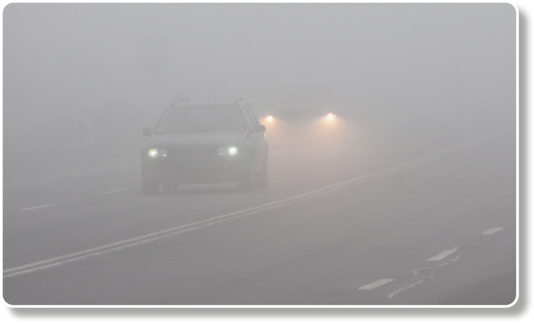 Fotografia de dois carros circulando em uma via com muita neblina. O carro da frente está com os faróis baixos, e o de trás, com faróis altos.