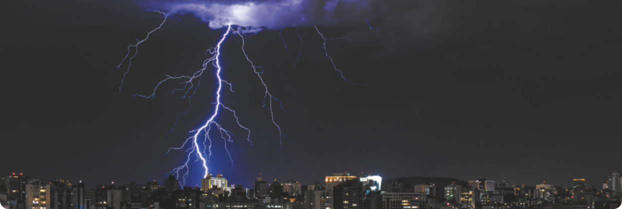 Fotografia aérea de uma cidade à noite. No céu, há uma nuvem da qual parte um relâmpago. O relâmpago é formado por linhas verticais e ramificadas, com aparência iluminada. A cidade aparece parcialmente iluminada por luzes artificiais.